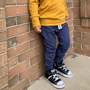 May include: A young boy wearing a mustard yellow long-sleeved shirt, blue jeans, and black high-top sneakers. He is standing in front of a brick wall.