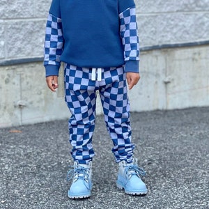 May include: A child wearing a blue sweatshirt with checkered sleeves and matching checkered pants. The outfit is paired with light blue boots and sunglasses. The background is a gray brick wall.