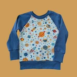 May include: A blue long-sleeved shirt with a raglan sleeve design. The shirt features a white background with a pattern of planets, stars, and rockets in various colors.