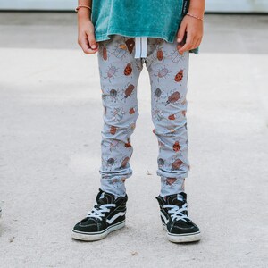 May include: A child wearing grey pants with a bug pattern and black high-top sneakers. The pants have a white drawstring.