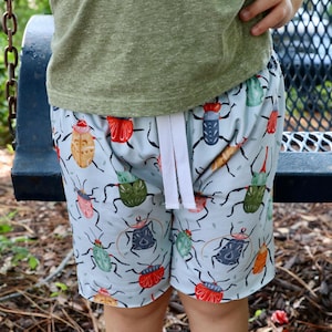 May include: Light blue shorts with a colorful insect pattern. The shorts have a white drawstring and are worn by a person. The insects are in shades of red, green, yellow, and blue. The shorts are made of a soft material.