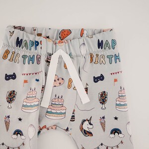 May include: Grey baby trousers with a white drawstring and a colourful print of birthday party items, including cake, balloons, and unicorns. The trousers have the text "Happy Birthday" printed on them.
