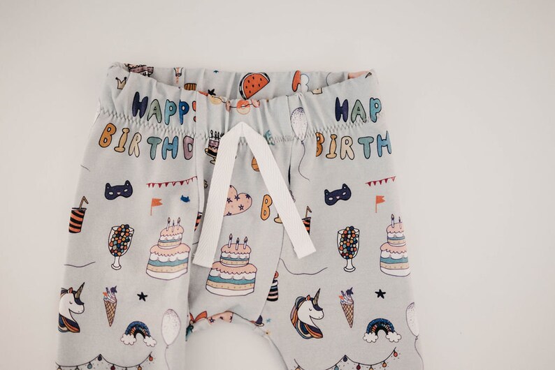 May include: Grey baby trousers with a white drawstring and a colourful print of birthday party items, including cake, balloons, and unicorns. The trousers have the text "Happy Birthday" printed on them.