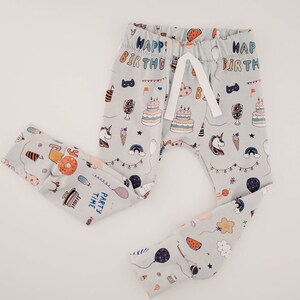 May include: Grey baby leggings with a birthday party theme. The leggings feature a variety of colourful illustrations, including birthday cakes, balloons, unicorns, and the text "Happy Birthday" and "Party Time".