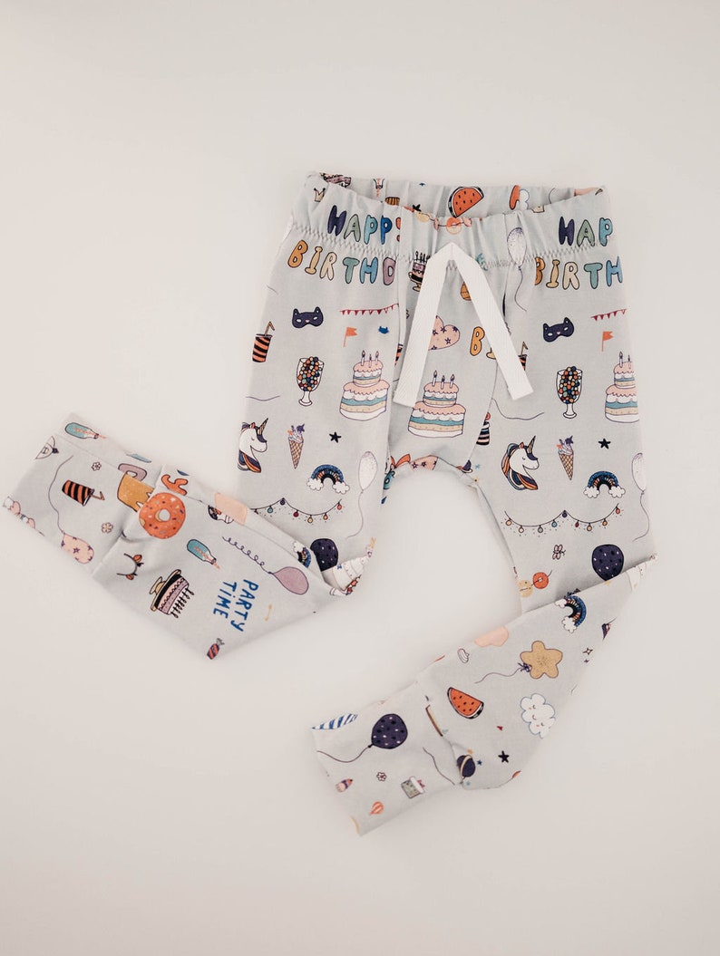 May include: Grey baby leggings with a birthday party theme. The leggings feature a variety of colourful illustrations, including birthday cakes, balloons, unicorns, and the text "Happy Birthday" and "Party Time".