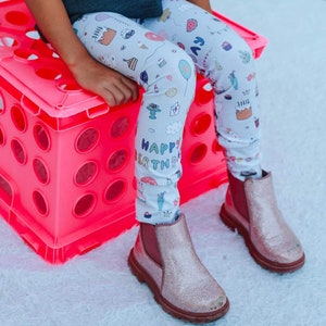 May include: A child wearing white leggings with a colourful birthday cake and party theme print. The leggings have the words "Happy Birthday" printed on them. The child is wearing pink glitter ankle boots.