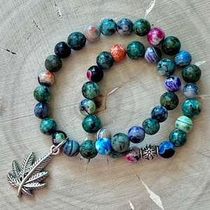 Puede incluir: Un brazalete de cuentas de ágata verde, azul y rosa con un dije de hoja de marihuana de plata. El brazalete está hecho de piedras naturales y tiene un aspecto único y terroso.