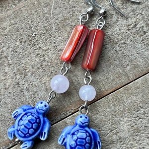 Puede incluir: Pendientes colgantes con abalorios de tortugas de cerámica azul, cuentas blanco-rosadas y cuentas cilíndricas de color marrón rojizo. Los pendientes tienen ganchos y alambres de conexión plateados. Sobre una superficie de madera envejecida.