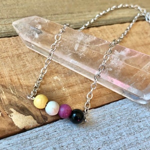 Op de afbeelding: Een zilveren ketting met een gele, witte, paarse en zwarte kralenbedel. De ketting ligt op een helder kristal.