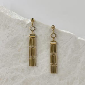 Puede incluir: Un par de pendientes colgantes de color dorado con un diseño de borla. Los pendientes están hechos de una cadena de pequeñas cuentas cilíndricas unidas.