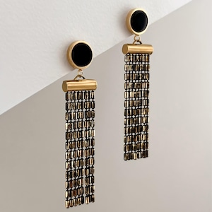Op de afbeelding: Een paar oorbellen met een ronde zwarte knop verbonden aan een goudkleurige cilindrische staaf. Aan de staaf hangen meerdere verticale strengen van kleine, metallic bronskleurige rechthoekige kralen die een franje-effect creëren.