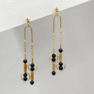 Puede incluir: Pendientes colgantes dorados con cuentas negras. Cada pendiente presenta una parte superior curvada, una cadena y una serie de cuentas negras y detalles dorados circulares y cilíndricos. Los pendientes son para orejas perforadas.