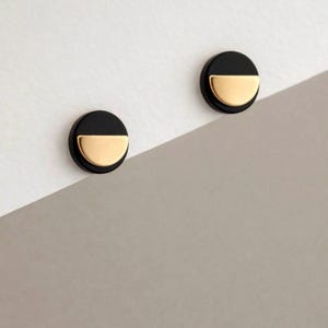 May include: A pair of circular stud earrings featuring a two-tone design. Each earring consists of a matte black circular base with a polished gold-tone semi-circular accent attached to the lower half, creating a distinct geometric contrast.