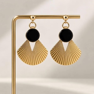 May include: A pair of gold-toned earrings featuring a circular black inlay at the top and a fan-shaped, pleated gold metal pendant suspended below. The earrings are attached to a gold-toned metal display stand with a horizontal bar.