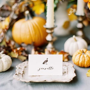 May include: A white place card with a small illustration of a turkey and the name "Jenrette" written in cursive. The place card is on a white plate with a decorative edge. The plate is on a gray surface with pumpkins and fall leaves.