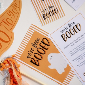 May include: Orange and white striped printable signs with the text "We've Been Booed" and "You've Been Booed" with a ghost illustration.  Instructions for a Halloween "Boo"ing game are included on a separate white card.