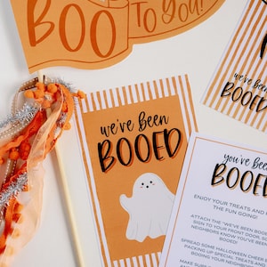 May include: Two orange and white striped cards with the text "we've been booed" and a ghost illustration. A third card with instructions for a "boo" game. The text reads "Enjoy your treats and the fun going! Attach the "we've been booed" sign to your front door, so neighbors know you've been booed! Spread some Halloween cheer and packing up special treats and booing your neighbors. Make sure to include this note and a "we've been booed" sign for them to use. Happy Halloween!"
