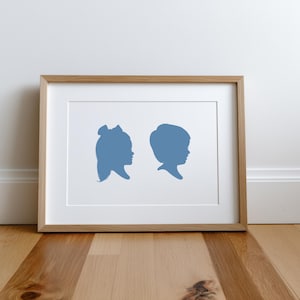 May include: Framed artwork featuring two blue silhouette portraits of children's profiles. The artwork is displayed in a wooden frame with a white mat, set against a white wall and hardwood floor.