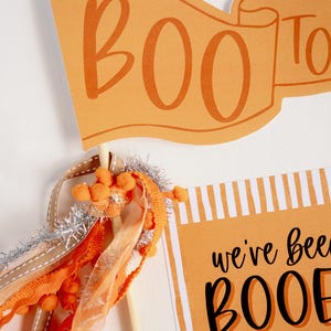 May include: Orange and white striped card with the text "we've been booed" and a cartoon ghost. A small orange banner with the text "BOO" is attached to a wooden stick with orange and silver ribbon.