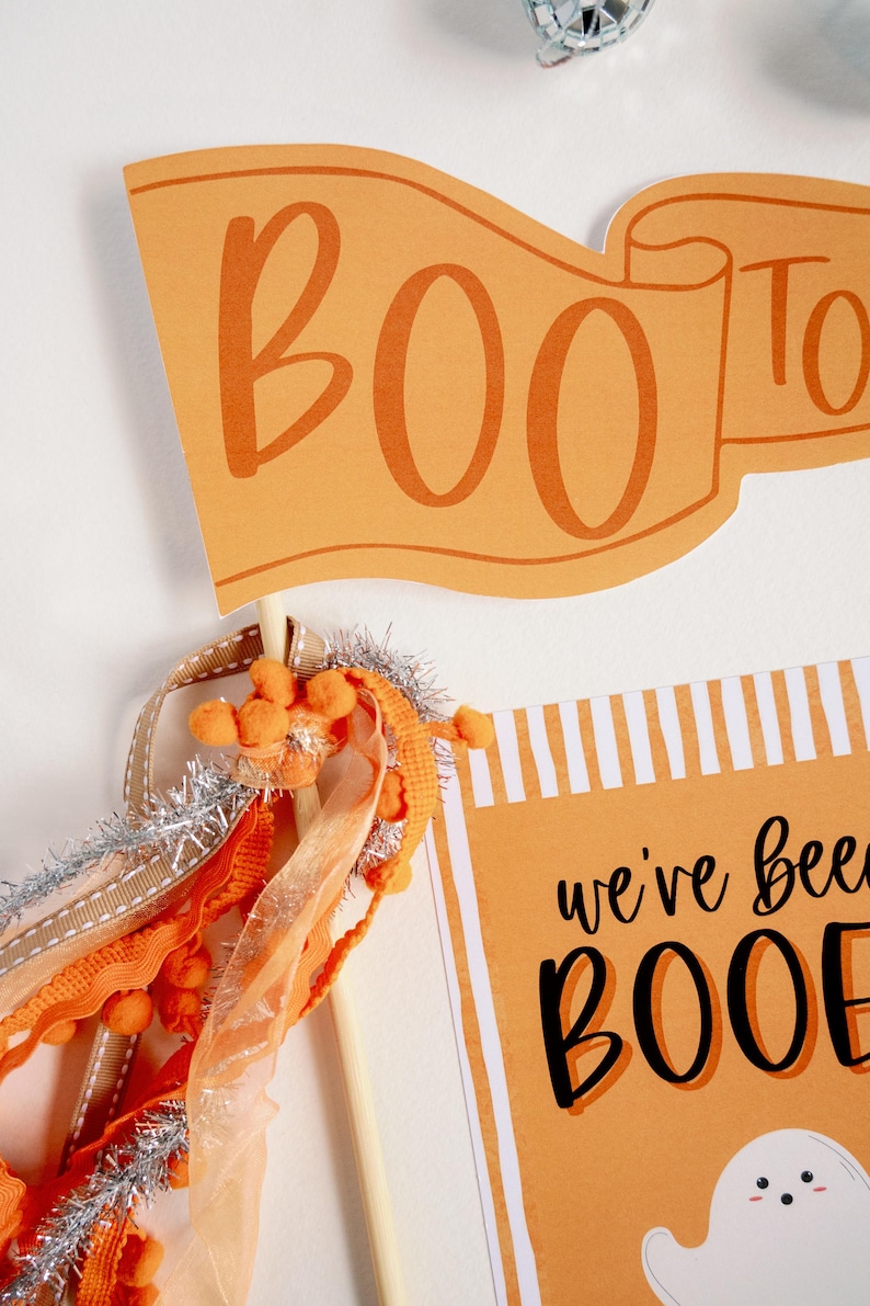 May include: Orange and white striped card with the text "we've been booed" and a cartoon ghost. A small orange banner with the text "BOO" is attached to a wooden stick with orange and silver ribbon.