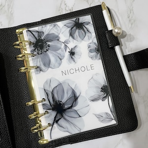 May include: Black leather planner with gold-colored rings, a floral insert, and a white pen with a pearl accent. The insert features a grayscale floral design and the name "NICHOLE". The pen is secured by a black leather loop.
