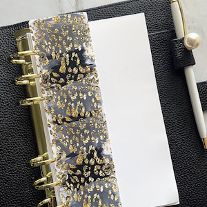 May include: Black leather planner with gold hardware, a white pen with a pearl accent, and a decorative divider. The divider features a gray and black watercolor design with gold glitter accents. The planner is open to reveal blank white pages.