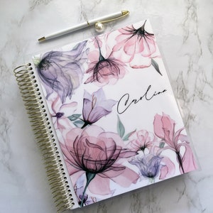 May include: A white planner with a clear plastic cover featuring a watercolor floral design in shades of pink, purple, and green. The planner has a gold spiral binding and the name "Carolina" is written in black cursive on the cover.