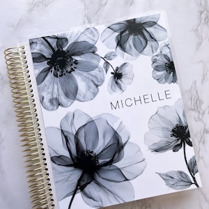 May include: A white planner with a gold spiral binding. The cover features a floral design with black and gray flowers. The name "Michelle" is printed in black at the bottom of the cover.