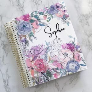 May include: A white spiral bound notebook with a floral design featuring pink, purple, and blue watercolor flowers. The name "Sophia" is written in black script on the front cover.