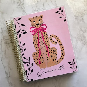 May include: A pink spiral-bound notebook featuring a cheetah illustration with a pink bow. The cheetah has black spots and is set against a pink background. Black leaf accents frame the design, and the name "Samantha" is written in cursive.