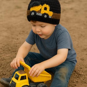 May include: A black knit beanie with a yellow and brown truck design and a dark blue pom-pom. The beanie is worn by a child playing with a yellow toy dump truck. The child is wearing a blue t-shirt and jeans.