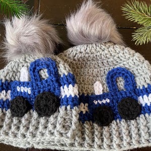 May include: Two gray crocheted hats with blue and white truck designs. Each hat has a gray faux fur pom pom on top.