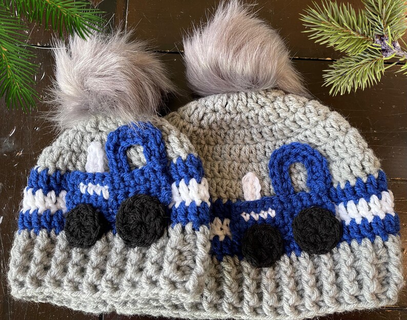 May include: Two gray crocheted hats with blue and white truck designs. Each hat has a gray faux fur pom pom on top.