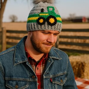 May include: A gray crocheted beanie with a green and yellow tractor design and a faux fur pom pom on top.