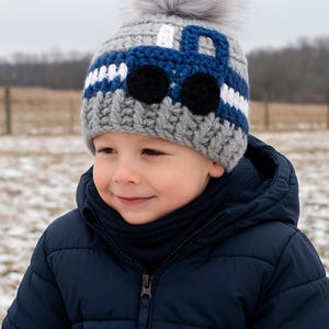 May include: A young child wearing a gray and blue knitted hat with a white pom-pom and a blue truck design. The hat has black eyes and a white stripe.