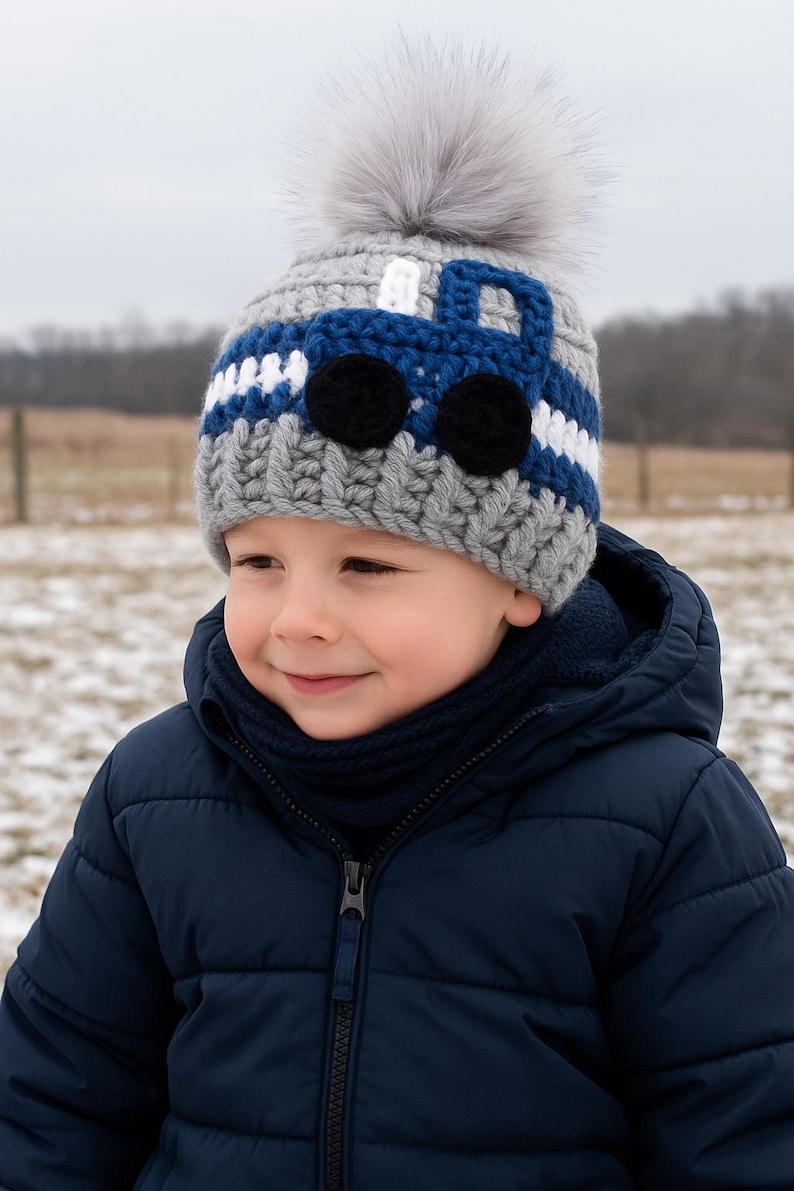 May include: A young child wearing a gray and blue knitted hat with a white pom-pom and a blue truck design. The hat has black eyes and a white stripe.