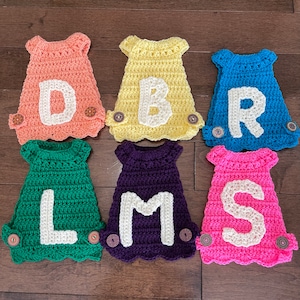 Peut inclure: Six ornements faits à la main en crochet en forme de robe de bébé, de couleurs variées : pêche, jaune, bleu, vert, violet et rose. Chaque robe comporte une lettre blanche et deux boutons décoratifs. Les lettres sont D, B, R, L, M et S.