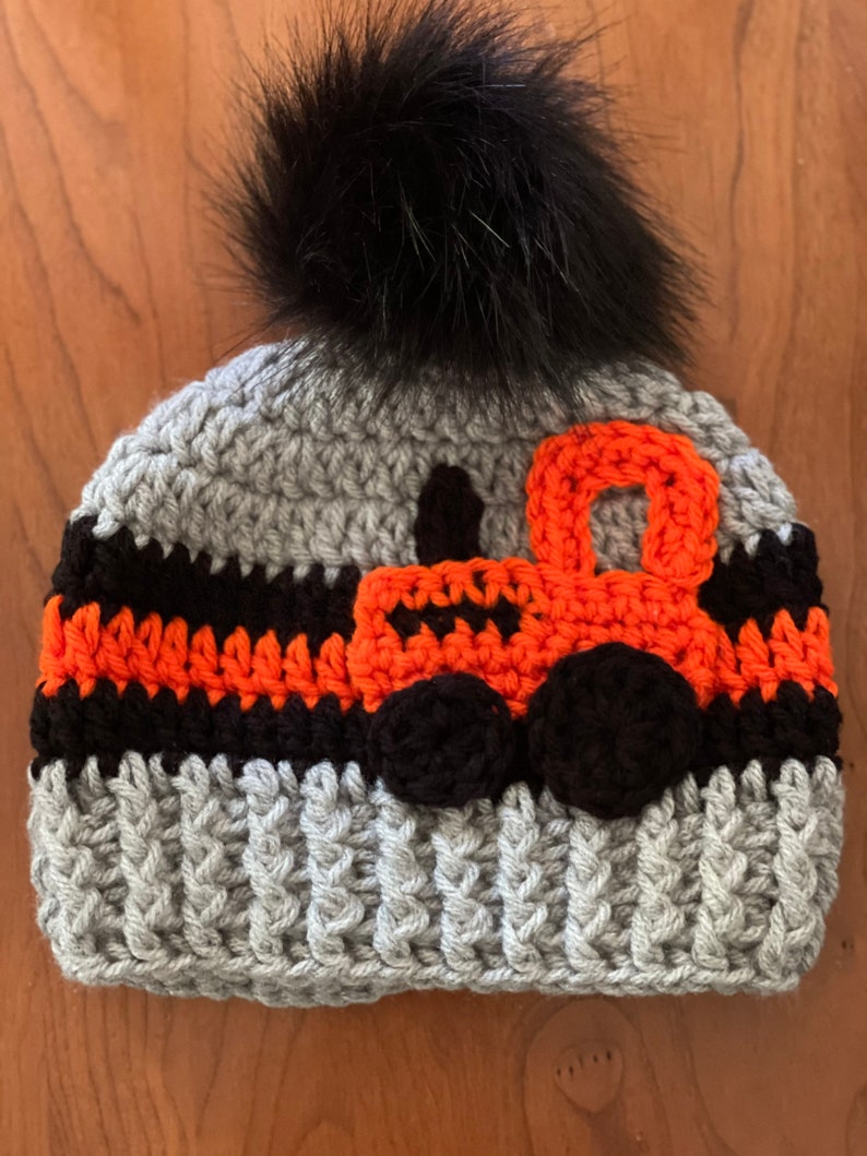 May include: Gray crocheted hat with a black pom pom and an orange and black crocheted tractor design.