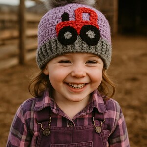 May include: A young child wearing a purple and gray crocheted hat with a red tractor applique. The hat has a fluffy gray pom pom on top. The child is smiling and wearing a purple plaid shirt and overalls.