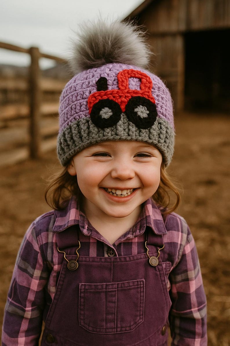May include: A young child wearing a purple and gray crocheted hat with a red tractor applique. The hat has a fluffy gray pom pom on top. The child is smiling and wearing a purple plaid shirt and overalls.