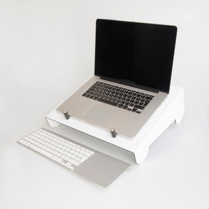 May include: A white laptop stand with a laptop on top of it. The laptop has a black screen and a silver keyboard. A white wireless keyboard with a silver trackpad is in front of the stand.