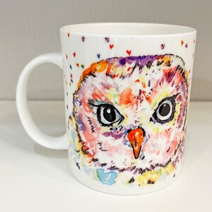 May include: A white ceramic mug featuring a colourful watercolour owl design. The owl has pink, orange, yellow and purple feathers. The background is white with small pink hearts.