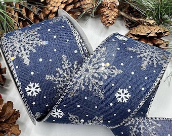Op de afbeelding: Een marineblauw lint met ijzerdraad, voorzien van een patroon van zilveren glinsterende sneeuwvlokken en kleine witte stippen. Het lint ligt tussen een decoratief arrangement van groene dennentakken, bruine dennenappels en rode bessen.