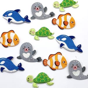 Könnte beinhalten: Eine Sammlung von zwölf bestickten Aufbügel-Patches mit verschiedenen Meerestieren, darunter blau-weiße Orcas, graue Robben mit weißen herzförmigen Bäuchen, orange-weiße Clownfische und grüne Meeresschildkröten mit gemusterten Panzern.
