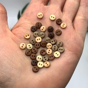 May include: A collection of small, two-hole buttons in brown, beige, and cream colours. The buttons are arranged in a pile on a person's hand.