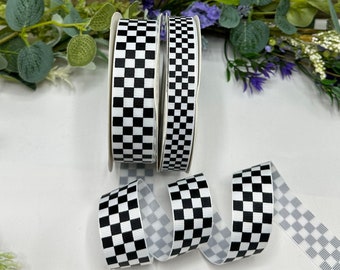 May include: Two rolls of black and white checkered ribbon. The ribbon is on a white surface with green and purple flowers in the background.