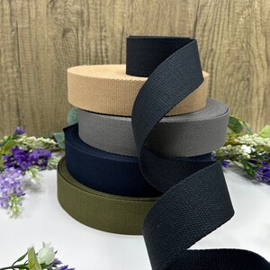 May include: A stack of five cotton webbing straps in different colors, including black, khaki, dark green, navy blue, and gray. The straps are rolled up and stacked on top of each other.