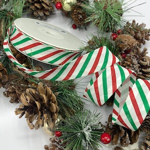 May include: A roll of striped ribbon with red, white, and green diagonal stripes. The ribbon is displayed with a Christmas wreath made of pinecones, evergreen branches, red berries, and gold ornaments. Festive holiday decor.