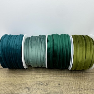 May include: Four spools of velvet ribbon arranged in a row, featuring a gradient of green shades including deep teal, muted sage, forest green, and olive. Each spool is wound with a continuous length of soft, textured fabric ribbon.