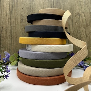 May include: A stack of colorful canvas webbing rolls in various colors, including black, tan, gray, navy, yellow, off-white, lavender, olive green, and rust. A length of tan webbing curls down the side. The rolls are stacked on a white surface.
